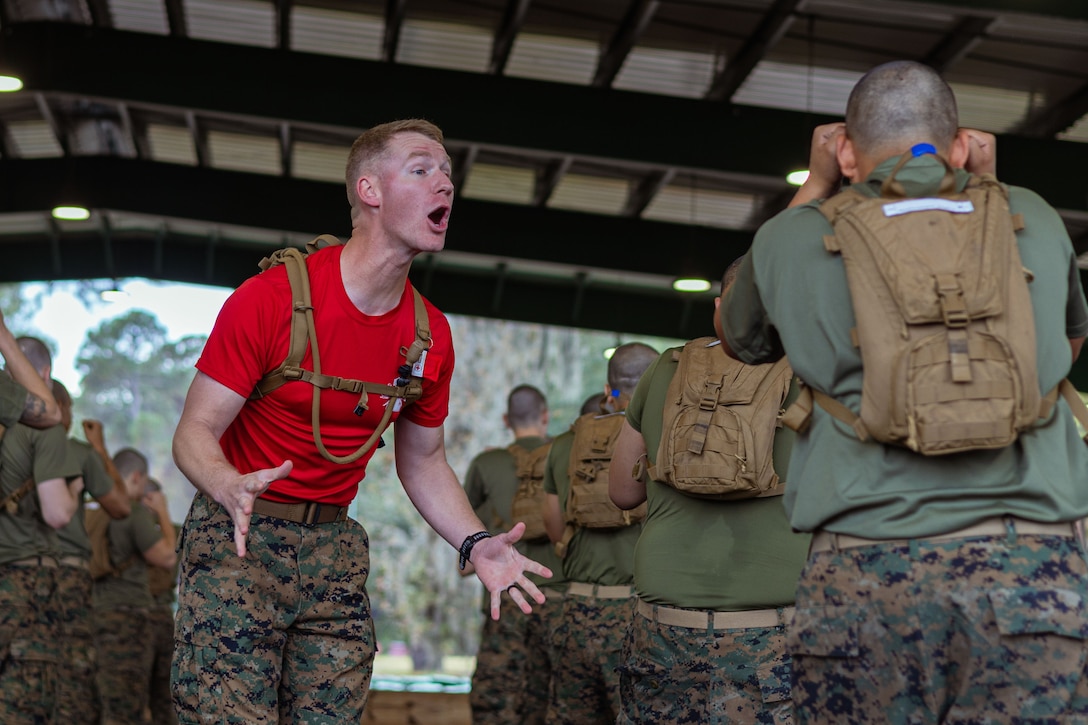 A Marine in a red T-shirt and camouflage pants yells toward a Marine recruit taking a boxing position, with other Marine recruits standing nearby in rows under a canopy outdoors during daytime.