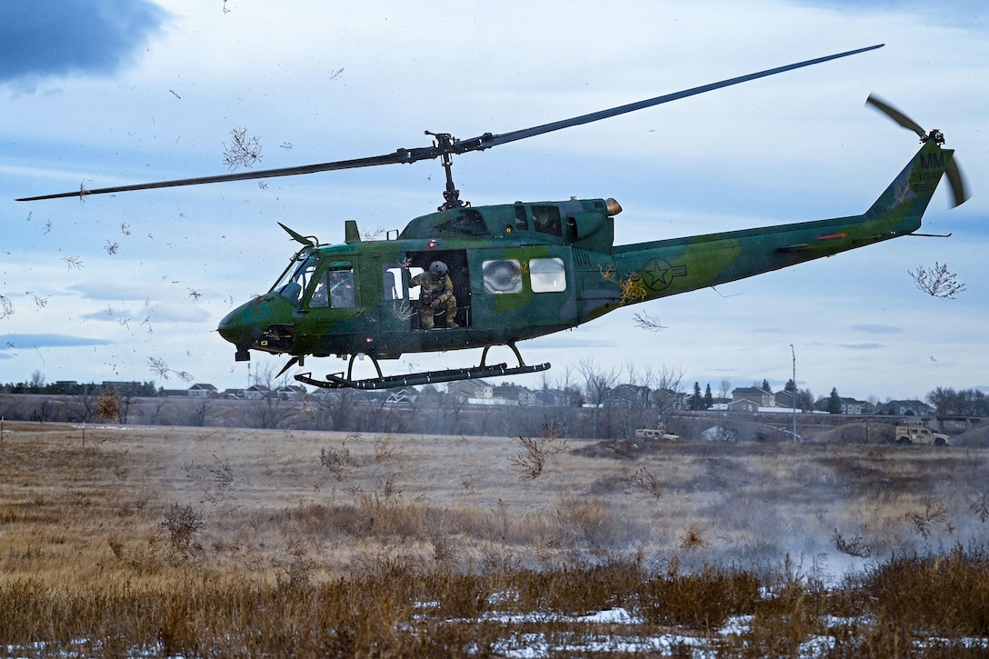 A green helicopter with a kneeling person looking backward out of an open door prepares to land in a field, kicking up vegetation from the ground during daytime.