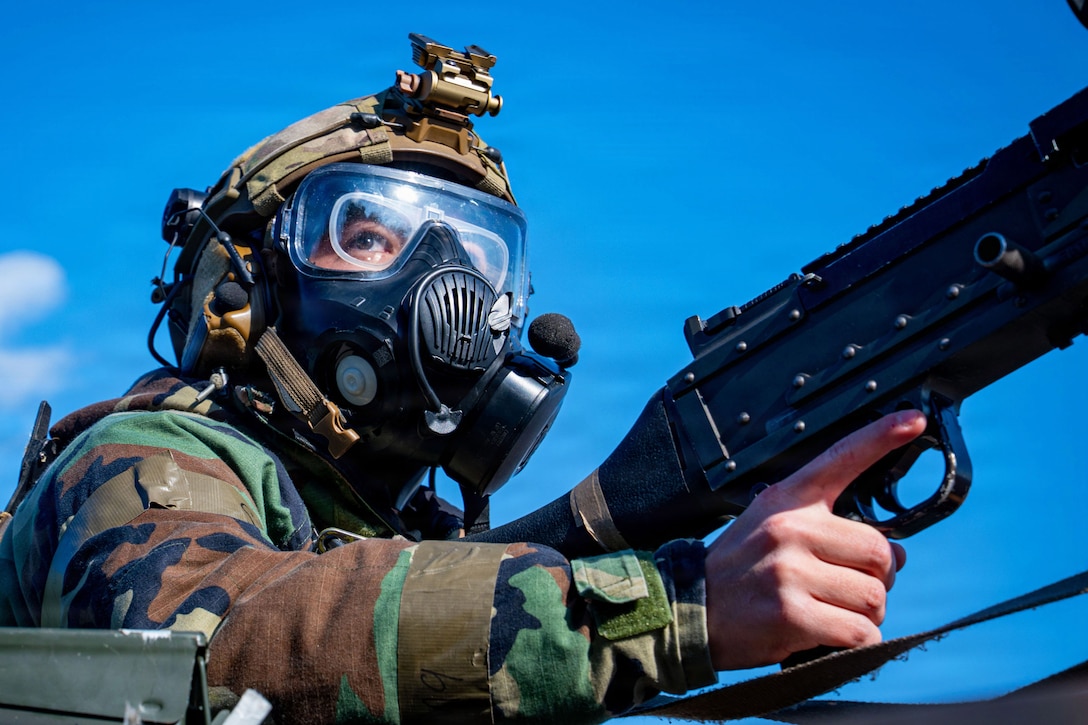 An airman wearing a gas mask and helmet holds a machine gun and looks into the distance during daytime, with a blue sky and cloud overhead.
