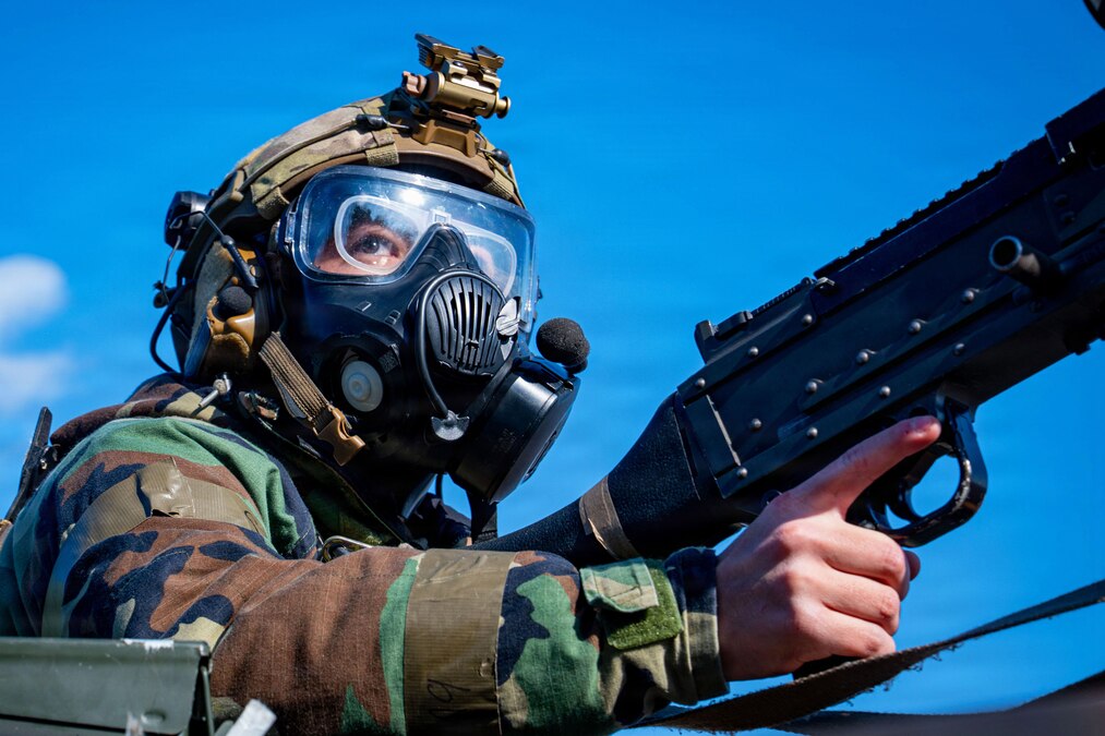 An airman wearing a gas mask and helmet holds a machine gun and looks into the distance during daytime, with a blue sky and cloud overhead.