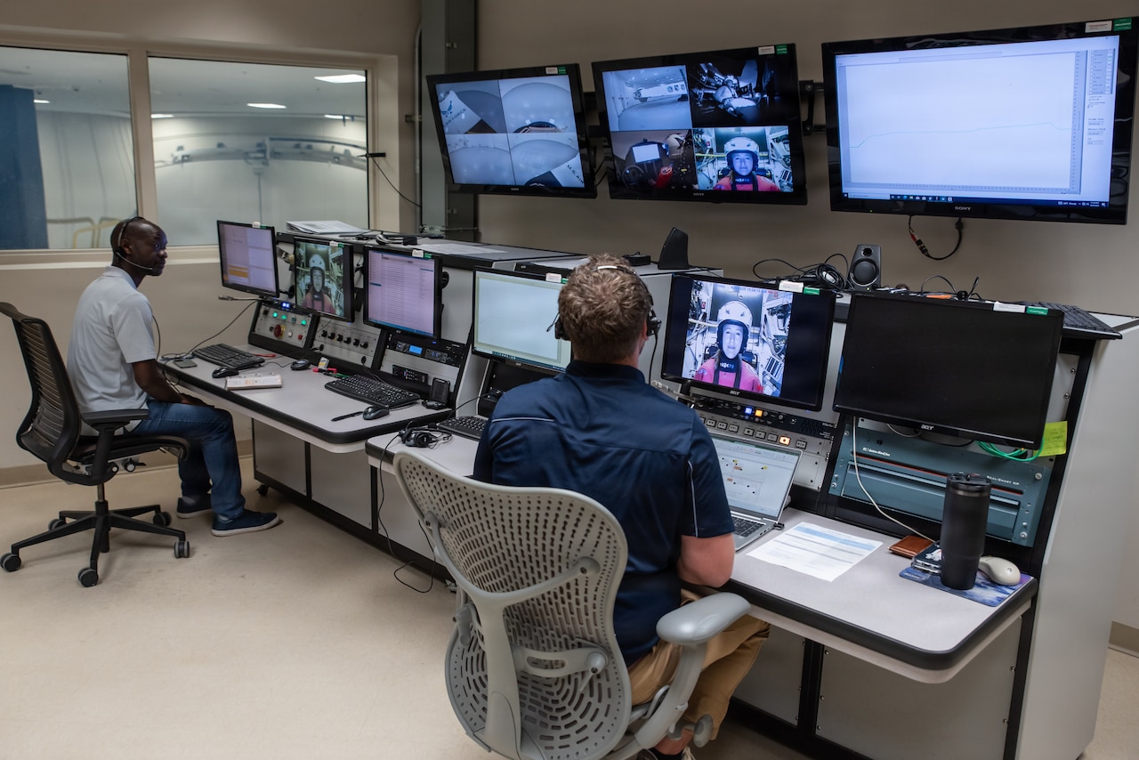 Two men wearing civilian attire and headsets sit in front of computer screens.