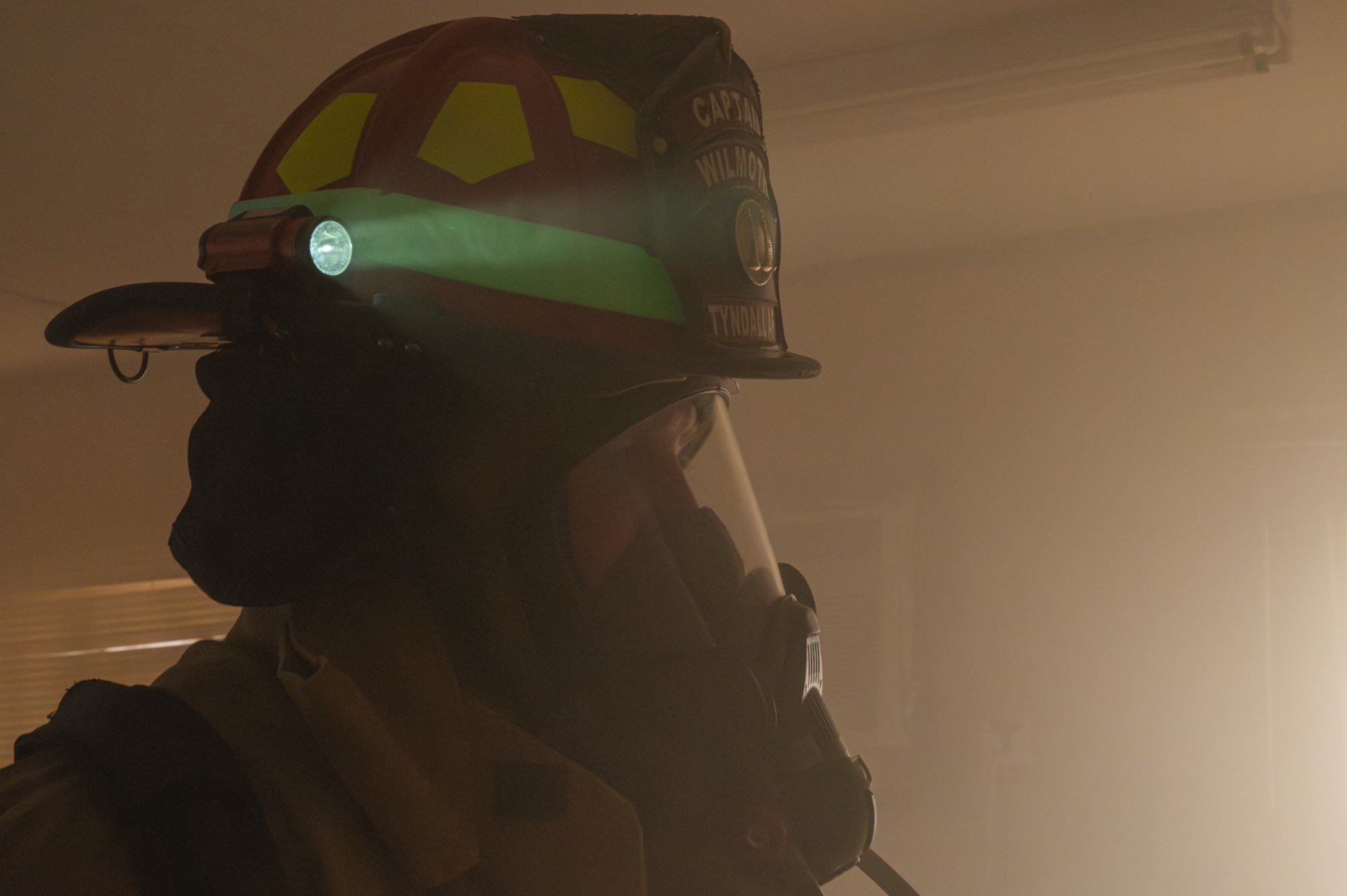 A firefighter, with his mask on, stands in a smoky room. A light attached to his helmet is illuminated.