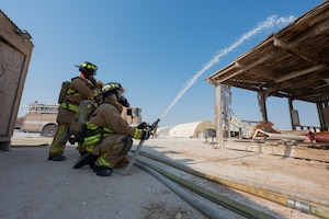 One fireman kneels and discharges their line toward a structure while another stands to the side