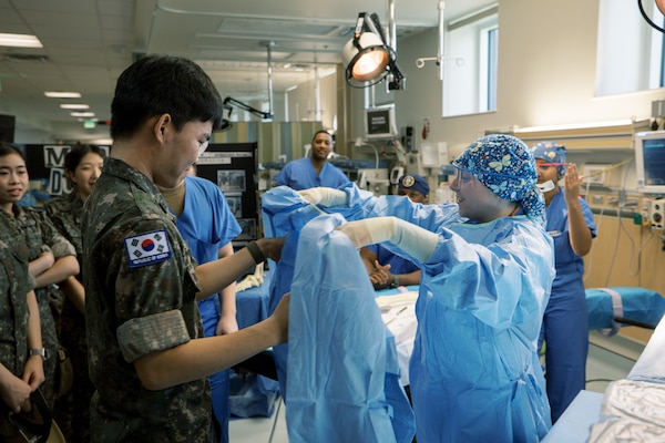 Hospital Corpsman 3rd Class Ashli Heaton, a surgical technologist, demonstrates the proper procedure for donning a surgical gown with a Republic of Korea (ROK) Military Nursing Academy cadet during a surgical skills rotation at U.S. Naval Hospital Guam, Nov. 14, 2025. The skills station introduced cadets to sterile techniques and operating room protocols as part of a hands-on tour of the military treatment facility's clinical departments. (U.S. Navy photo by Hospital Corpsman 1st Class Clien Lester Guico.)