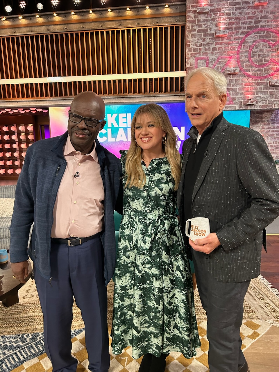 Retired Special Agent Leon Carroll on the left, singer and talk show host Kelly Clarkson in the middle, and actor Mark Harmon on the right stand together on the set of the Kelly Clarkson show.