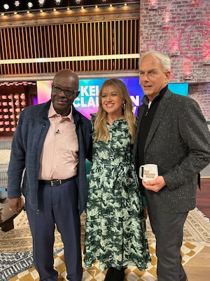 Retired Special Agent Leon Carroll on the left, singer and talk show host Kelly Clarkson in the middle, and actor Mark Harmon on the right stand together on the set of the Kelly Clarkson show.
