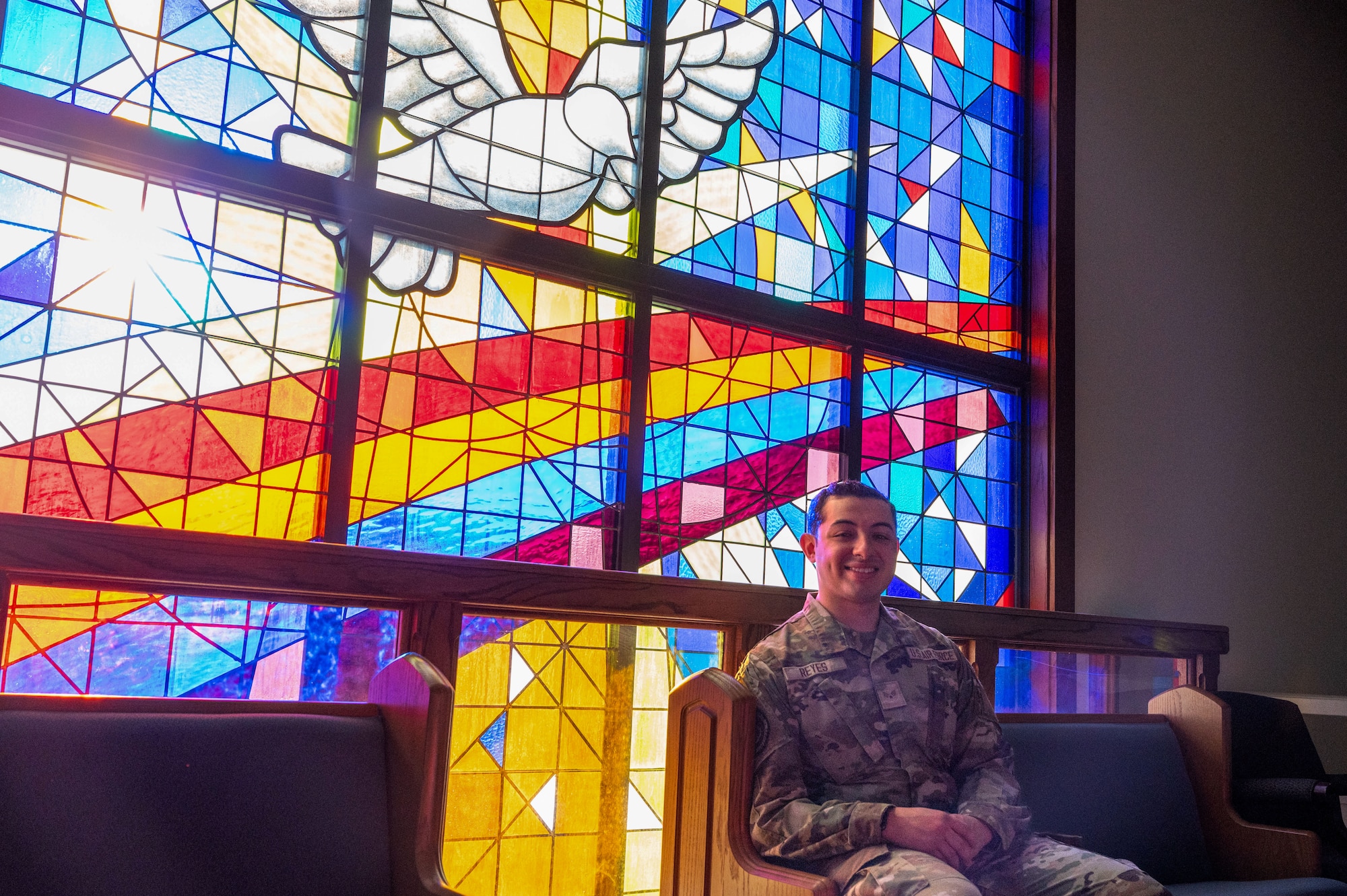 U.S. Air Force Senior Airman Joshua Reyes, 97th Air Mobility Wing Chapel Office religious affairs airman, sits by a stained glass window at Altus Air Force Base, Oklahoma, Sept. 29, 2025. Reyes takes pride in his everyday work at the base chapel. (U.S. Air Force photo by Tech. Sgt. Hailey Haux)