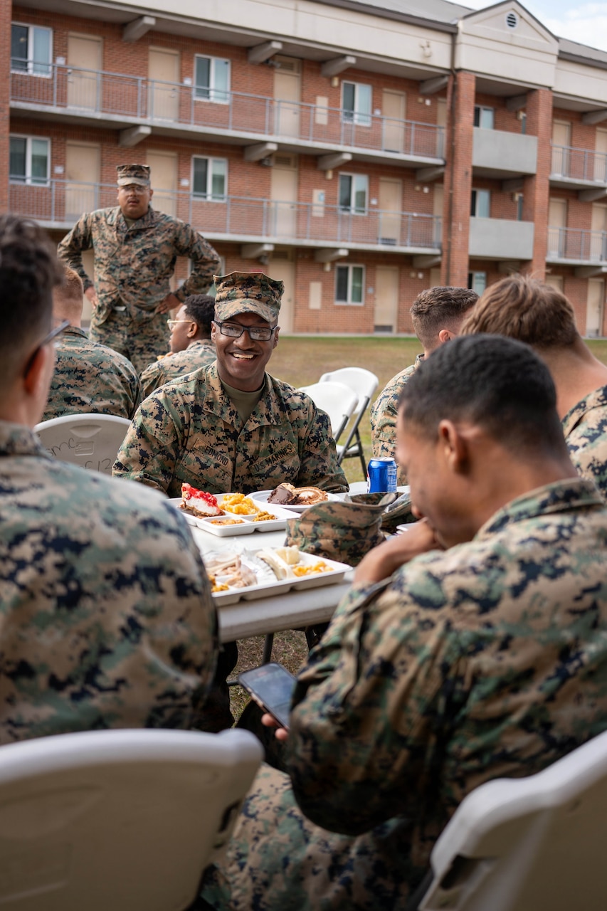 Troops in camouflage uniforms are seated at a table, eating outside in front of a building.
