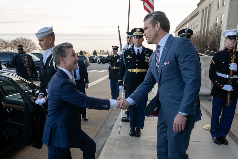 Two people wearing suits shake hands outdoors, with service members and a vehicle in the background during daytime.