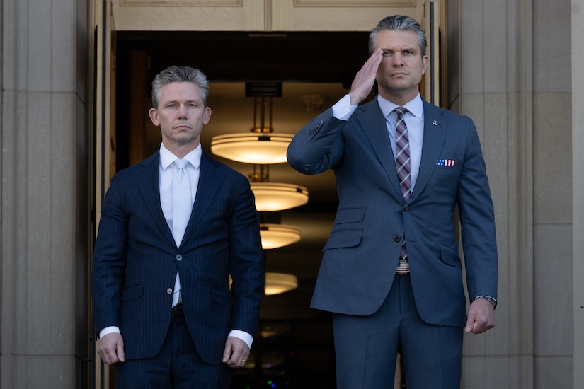 Two people wearing suits stand outdoors, one saluting, at the entrance to a building during daytime.