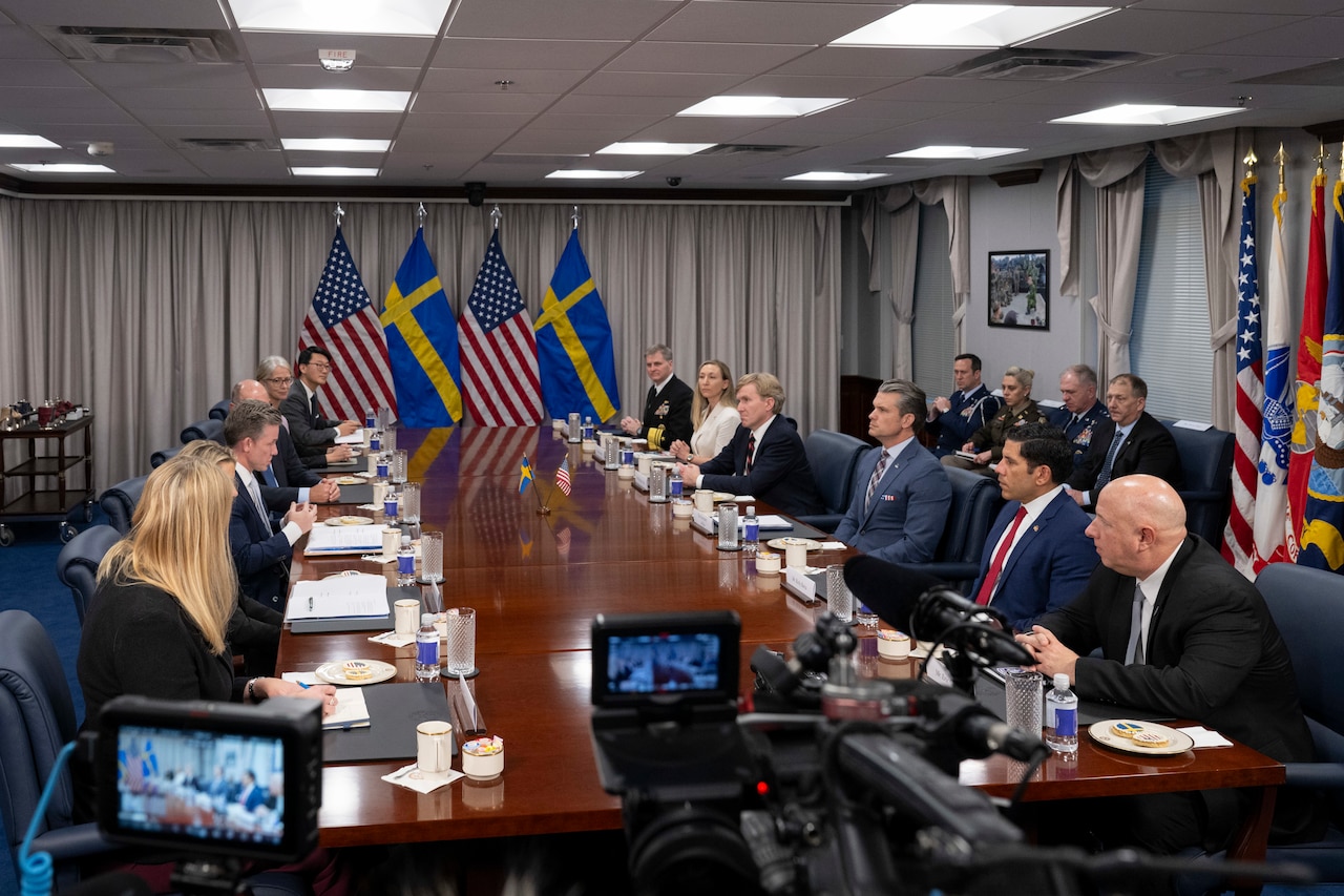 People in business attire and military uniforms sit around a large table in a conference room indoors.