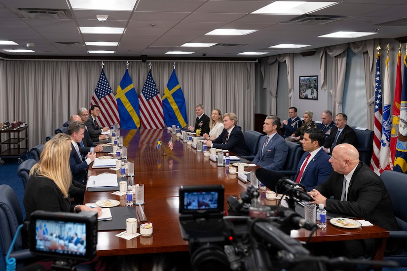 People in business attire and military uniforms sit around a large table in a conference room indoors.