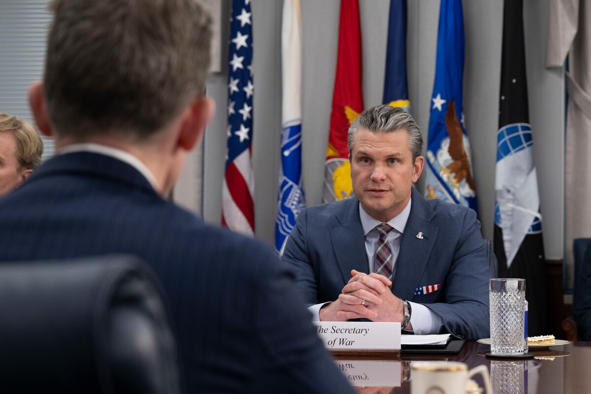Two people wearing suits and sitting across from each other at a table indoors speak, with flags drooped on poles in the background.