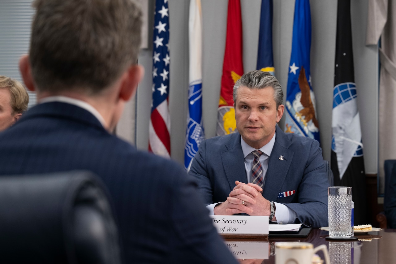 Two people wearing suits and sitting across from each other at a table indoors speak, with flags drooped on poles in the background.
