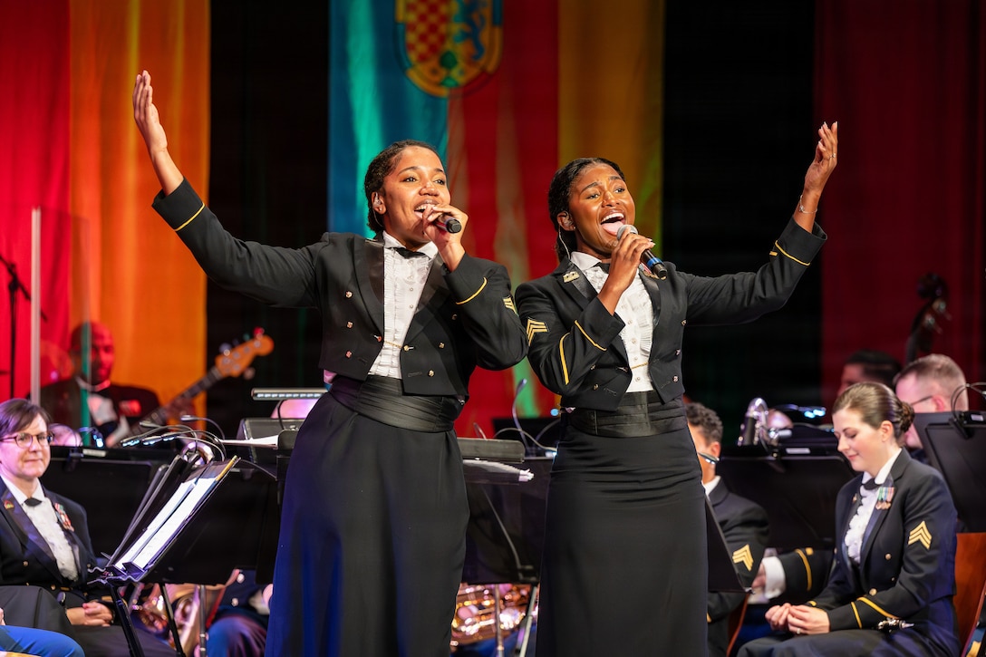 Two soldiers hold microphones and sing as other soldiers with musical instruments sit in the background in a stage-like setting indoors.