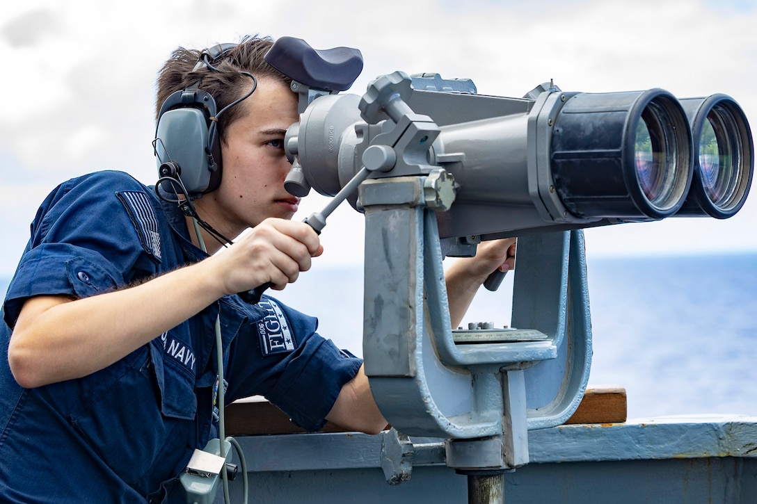 A sailor looks through large binoculars mounted on a ship during daytime, with a large body of water in the background.