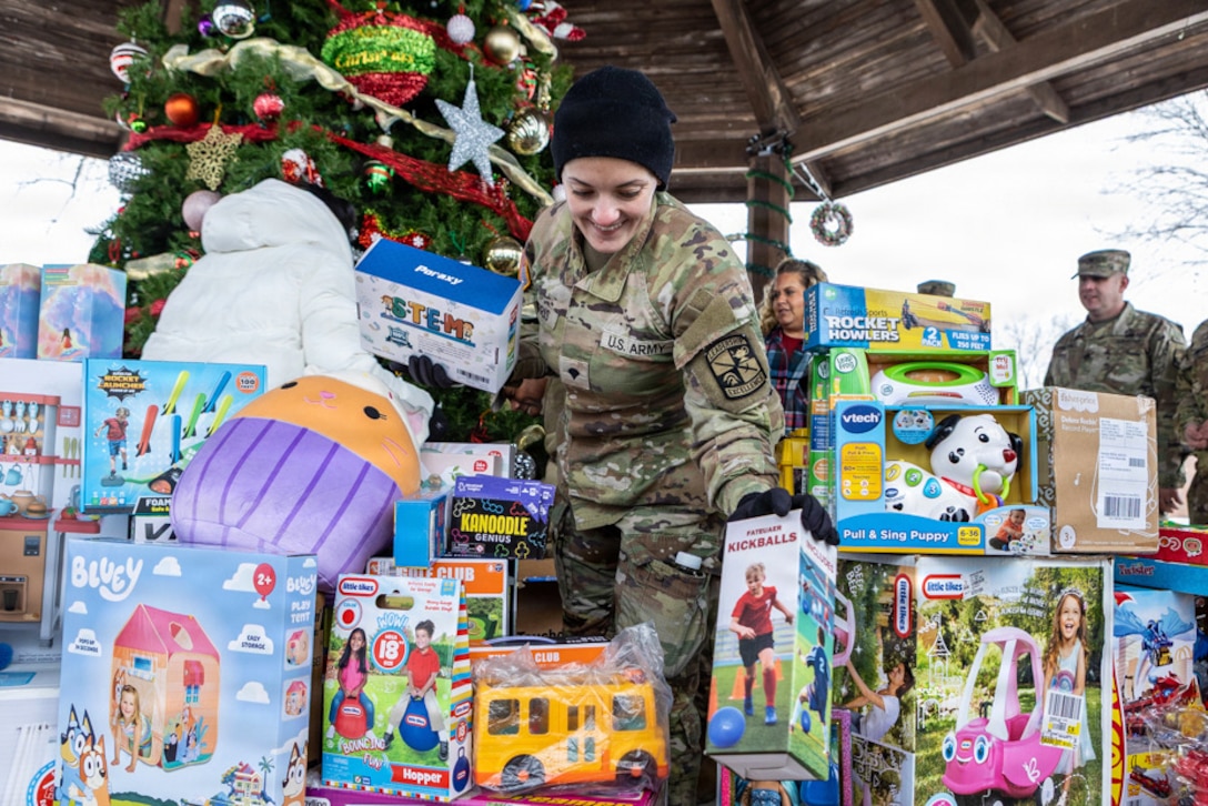 A soldier places boxes of toys in a pile of other toys near a tree under a wooden roof outdoors, as other soldiers and people stand in the background.