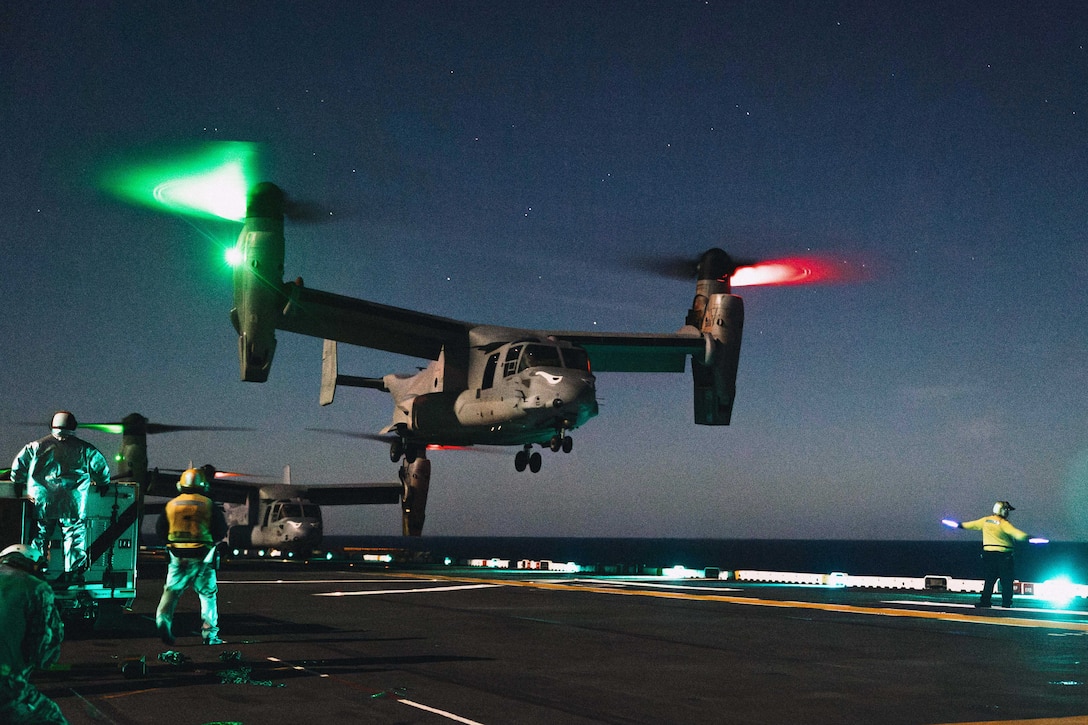 A tiltrotor aircraft with green and red lights on its rotors lands on the deck of a ship, as service members stand nearby under a dark sky.