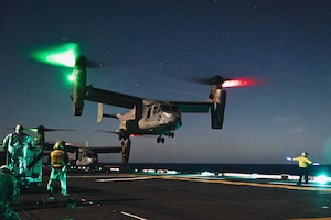 A tiltrotor aircraft with green and red lights on its rotors lands on the deck of a ship, as service members stand nearby under a dark sky.