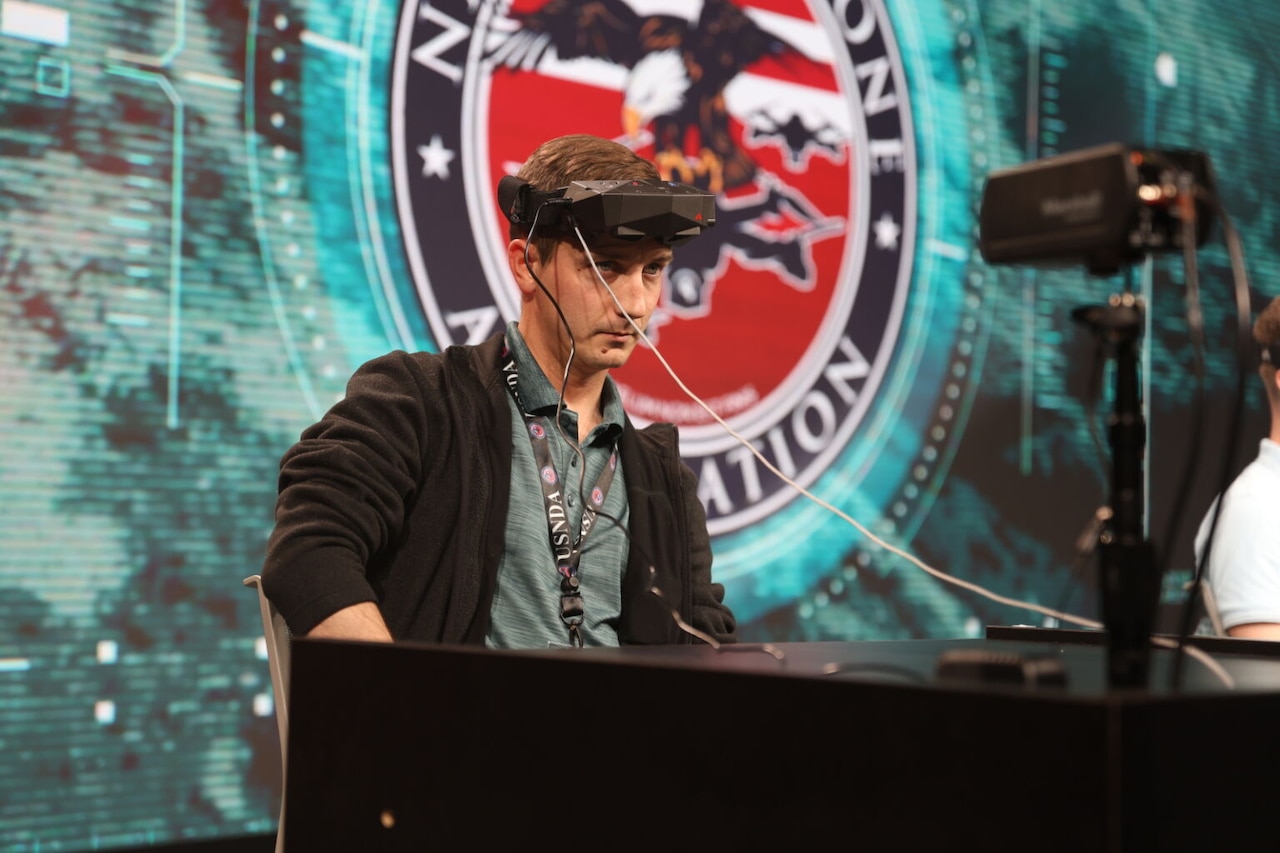 A man in casual attire wears a virtual reality headset on his head and stands at a lectern.