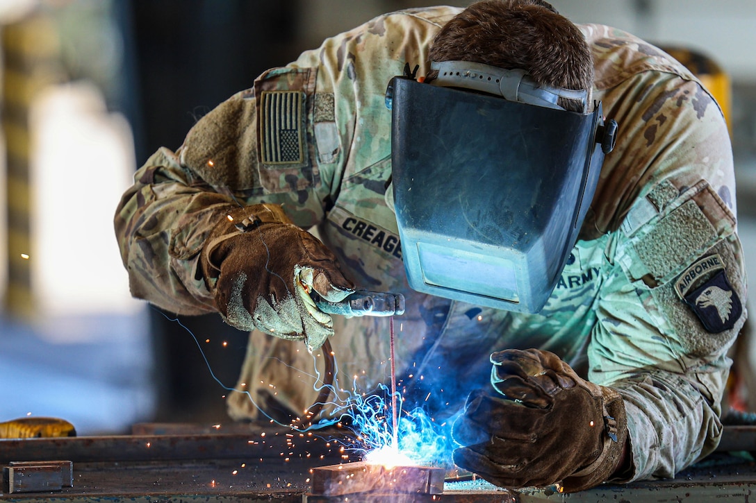A soldier wearing heavy gloves and a face shield welds a metal post in a shop-like setting.