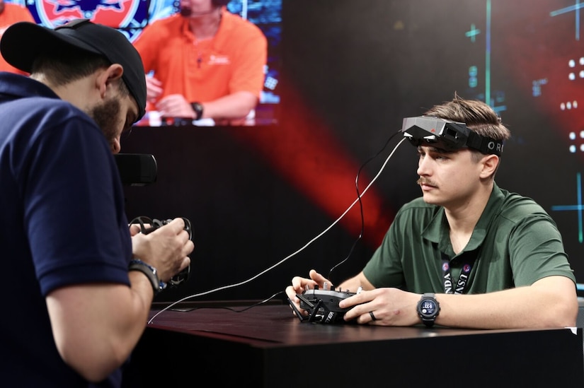 A man in casual attire wears a virtual reality headset and sits at a table observing another man as he holds a remote control.