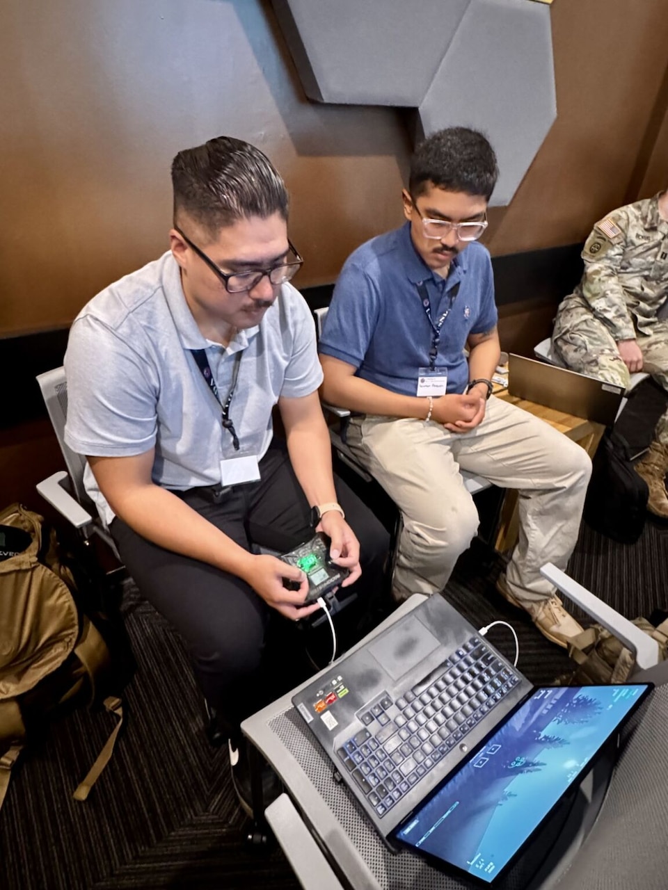 Two men in casual attire sit at a laptop; one is holding a remote control.