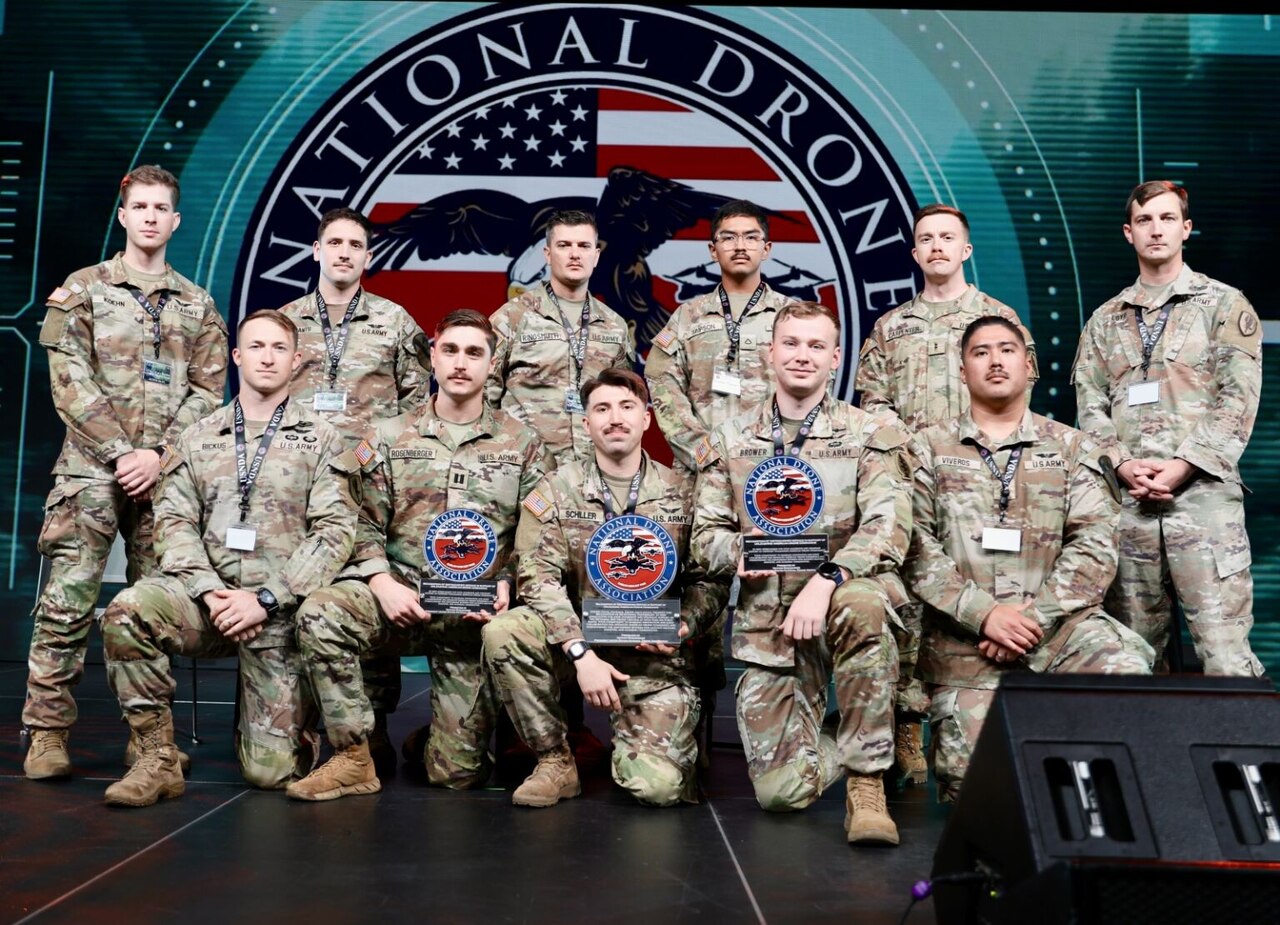 A group of 11 men in camouflage military uniforms pose for a photo; three in the front hold trophies.