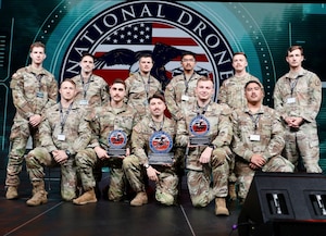 A group of 11 men in camouflage military uniforms pose for a photo; three in the front hold trophies.