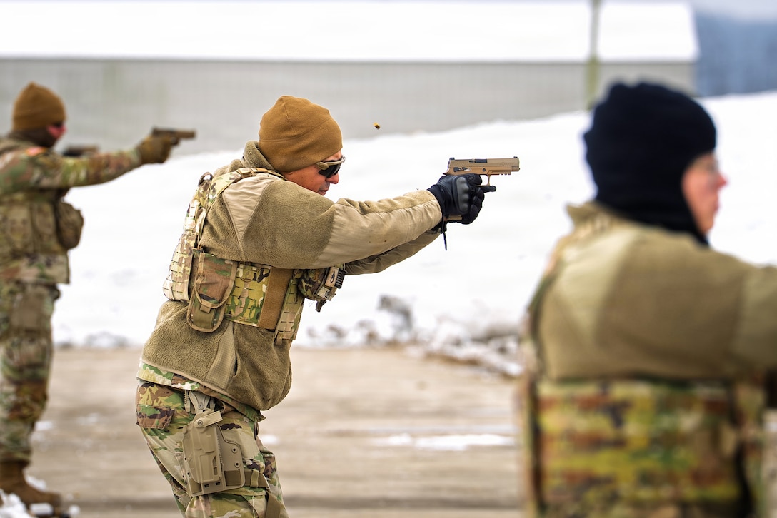 A guardsman stands in a row with other guardsmen outdoors and fires a pistol, with snow and a building in the background.