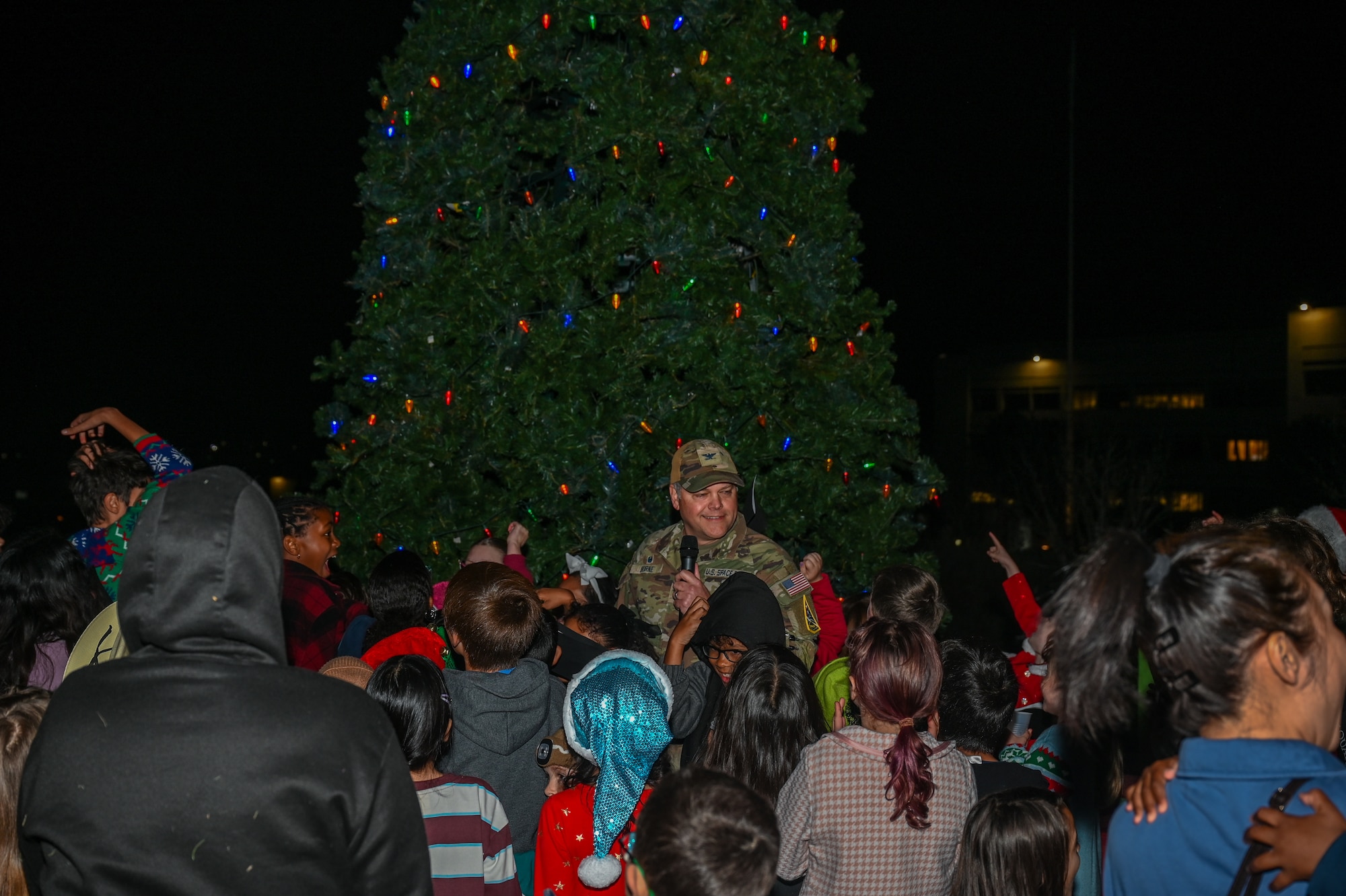 U.S. Space Force Col. James T. Horne III, Space Launch Delta 30 commander, stands in front of a Christmas tree outside. He holds a microphone and is speaking to a group of people standing in front of him.