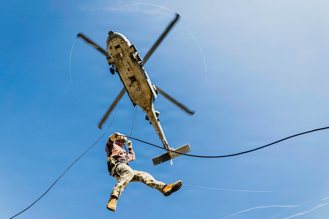 A sailor rappels from a flying helicopter with a rope during daytime, with a blue sky overhead.