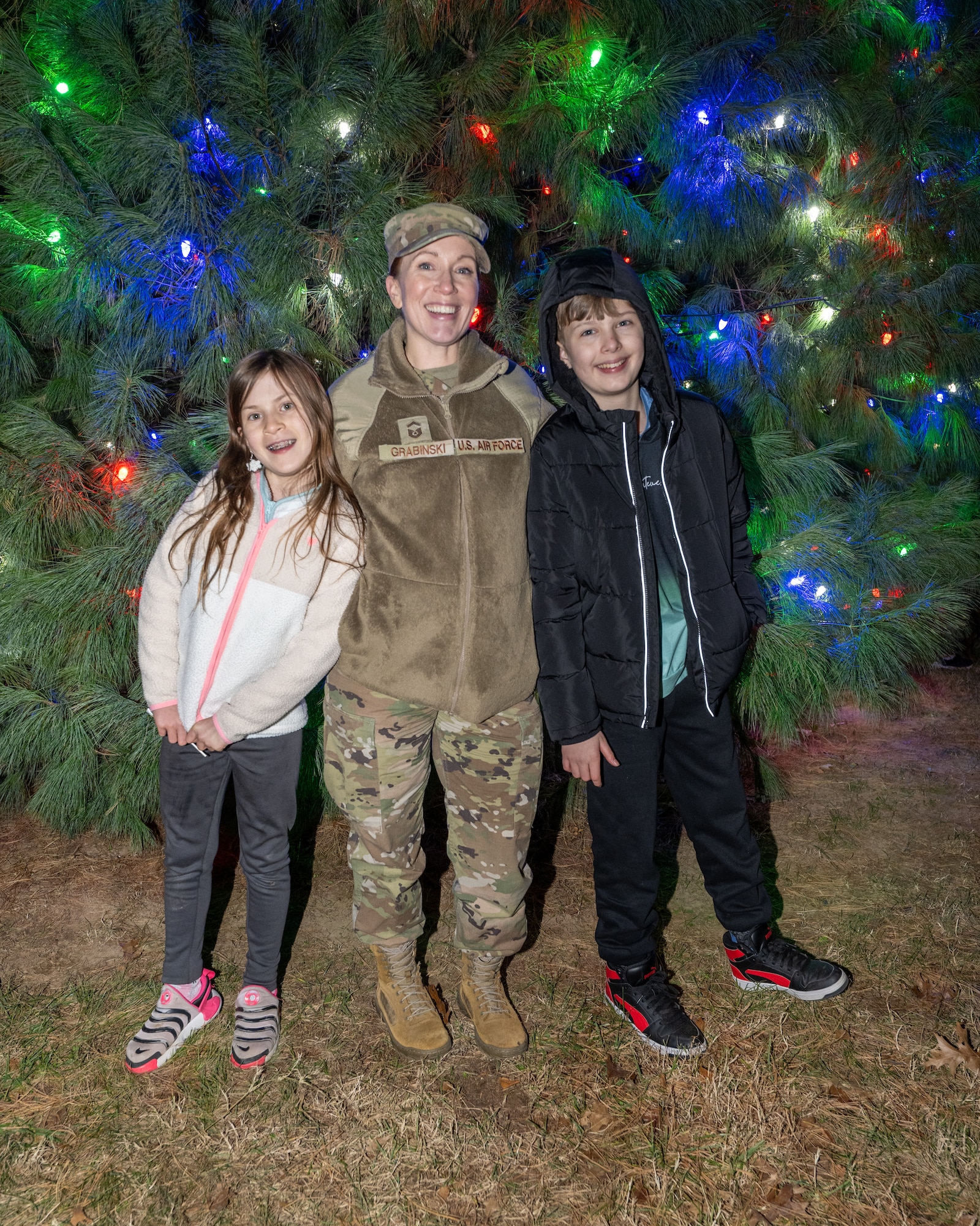 U.S. Air Force Senior Master Sgt. Jessica Grabinski, 436th Communication Squadron senior enlisted leader, and her children stand in front of the newly lit Christmas tree at the annual holiday parade at Dover Air Force Base, Delaware, Dec. 1, 2025. Each year, the family of a deployed Dover Airman is chosen to flip the switch turning on the lights for a crowd of spectators. (U.S. Air Force photo by Mauricio Campino)