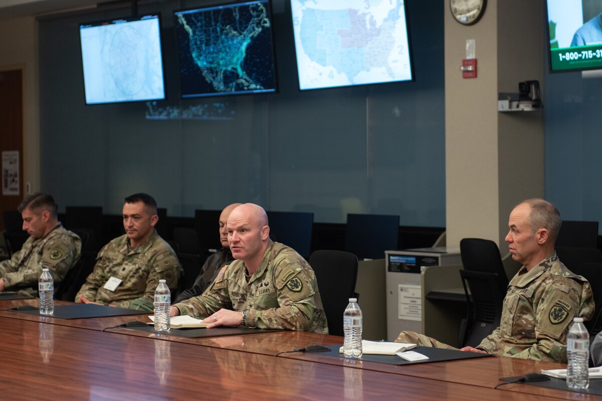 A man dressed in a military camouflage uniform seated at the center of a table speaks to other men seated around him. The others are also dressed in military camouflage uniforms.