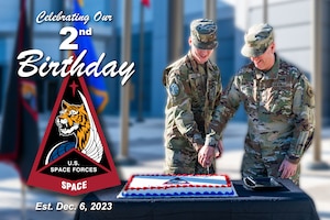 Two military members cut a cake in a courtyard with flags behind them.