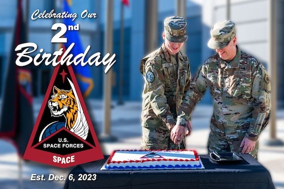 Two military members cut a cake in a courtyard with flags behind them.
