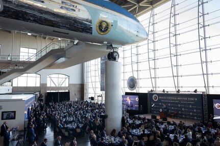 A view looking down on a large crowd of people sitting at tables in a ballroom, observing a man in a business suit speaking from behind a lectern on a stage. Above the people is a static display of a large passenger aircraft.