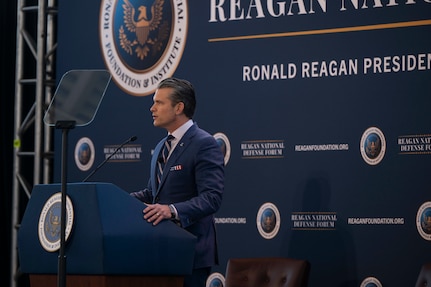 A man in a business suit stands behind a lectern on a stage, speaking into a microphone while reading from a teleprompter.