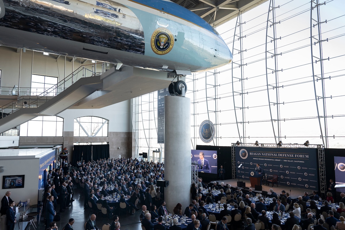 A view looking down on a large crowd of people sitting at tables in a ballroom, observing a man in a business suit speaking from behind a lectern on a stage. Above the people is a static display of a large passenger aircraft.