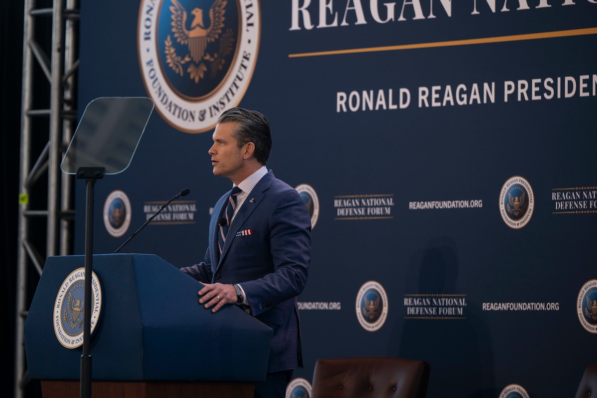 A man in a business suit stands behind a lectern on a stage, speaking into a microphone while reading from a teleprompter.