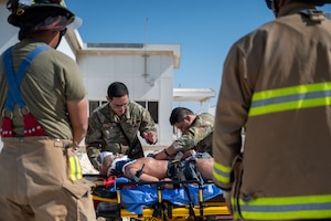 Two Airmen treat a dummy patient lying on a stretcher while two firefighters in the foreground watch