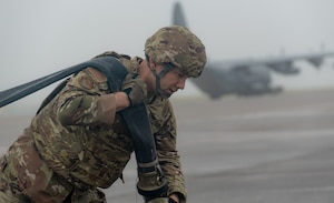 An Airman carries a hose across a runway with an aircraft parked behind him in the distance.