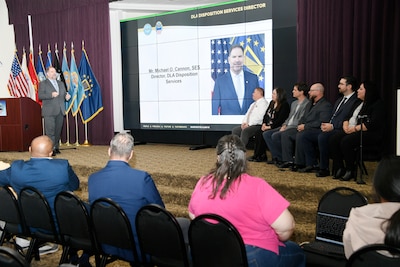 one person on stage talking, six people on the same stage looking at the speaker.