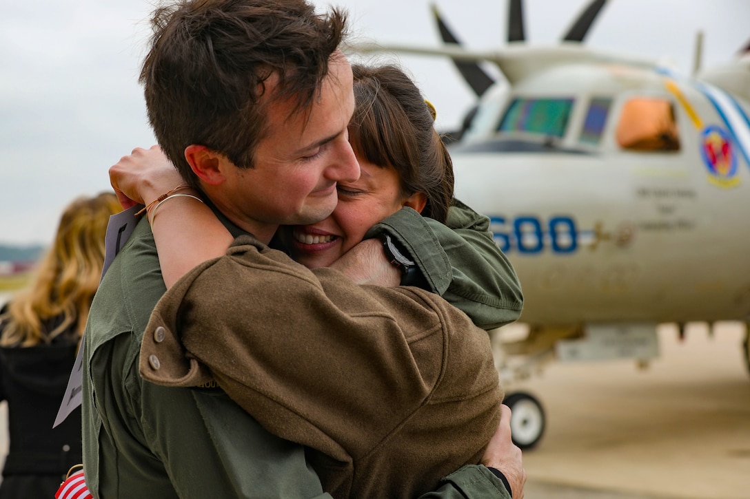 A sailor hugs a loved one as an aircraft sits to the right in the background.