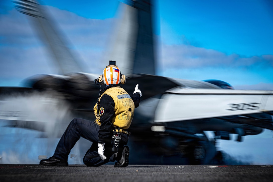 A kneeling sailor signals to a blurred aircraft as it swiftly launches from a ship at sea.