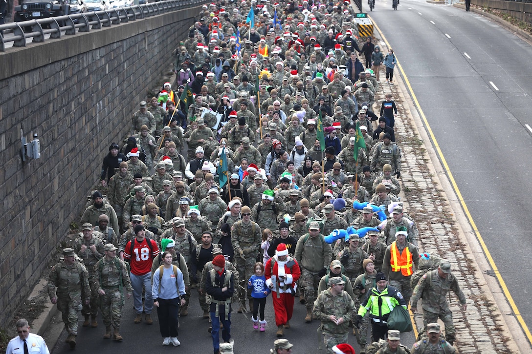 Dozens of service members wearing Santa hats and carrying backpacks march through a city as seen from above.