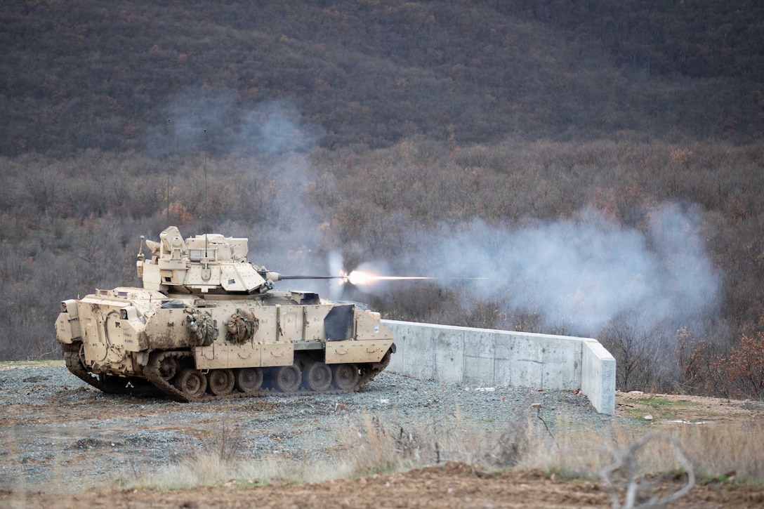 A tank fires toward leafless trees as smoke blows in the air.