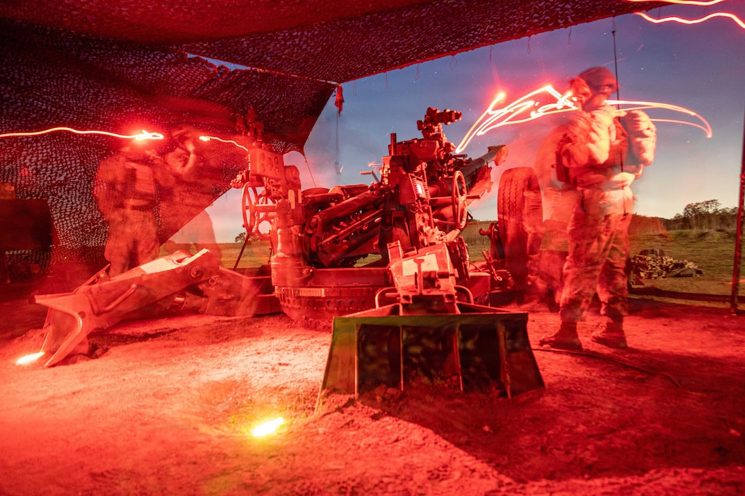 Marines fire a weapon toward a blue, starry sky under a makeshift shelter illuminated by red streaks.