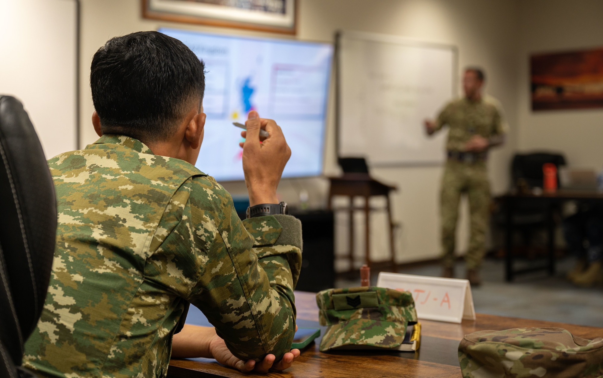 An Indonesian service member sits leaning forward looking at a presentation as they listen to a course instructor in a conference room.