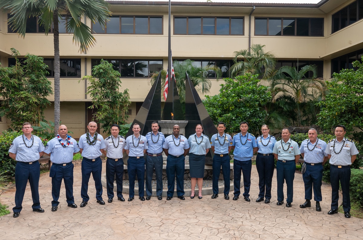 A group of service members stand in a horizontal line in front of the memorial in the Courtyard of Heroes, where an eternal flame is lit and flickering behind them.